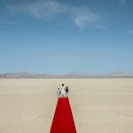 Image: A woman and two children stand in the desert, a red carpet spread behind them.