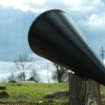 A black megaphone rests on a piece of wood with green pastures and blue skies in the background
