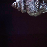 Image: Cropped photograph of a dancer on a dark stage who is lit from behind. A large silver seashell is suspended above her on stage.