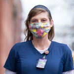 Image: A nurse with long brown hair poses on a New York City sidewalk wearing a headband, mask, scrubs, and an ID card. The background is blurred and they stand smiling and in focus in the center of the frame.