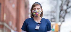 Image: A nurse with long brown hair poses on a New York City sidewalk wearing a headband, mask, scrubs, and an ID card. The background is blurred and they stand smiling and in focus in the center of the frame.