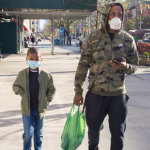Image: A man wearing a camouflage hoodie, athletic pants, and a mask carries a green plastic shopping bag and cell phone while walking on a New York City sidewalk in Harlem. His son walks beside him, also wearing a mask.