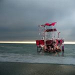 Photograph of mixed media sculpture that resembles a red raft on the sea and overcast skies