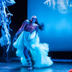 Image: Cropped photograph of a blue-lit dance performance. Two dancers, a woman and a man, appear on stage with jellyfish dangling from above. They are caught mid-movement against a screen with a seashell on it.