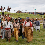 Image: A group of Black reenactors are pictured walking on grass and along a highway. They wear period dress from the early 1800s and carry weapons; some on higher ground are on horseback. A man in front carries a blue flag with a white emblem on it.