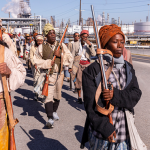 Image: A group of Black reenactors are pictured walking along a highway with what look to be oil refineries in the background. They wear period dress from the early 1800s and carry rifles.