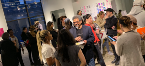 Image: A social gathering of artists participating in NYFA's Immigrant Artist Mentoring Program: Newark. It appears to be a mixer event, as people mingle in a large, gallery-like setting and hold drinks and plates of food.