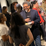 Image: A social gathering of artists participating in NYFA's Immigrant Artist Mentoring Program: Newark. It appears to be a mixer event, as people mingle in a large, gallery-like setting and hold drinks and plates of food.