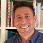 Image: A man smiles broadly into the camera, pictured from the shoulders up against what bookshelves. He wears a blue button-down shirt with small white dot print and has dark hair.