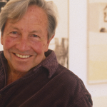 Image: A man, Robert Rauschenberg, is sitting in a white gallery-like setting in front of his own artworks. His hands are clasped together as he smiles into the camera.