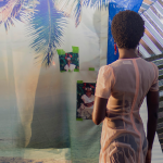 Image Detail: A color photograph of a figure standing in a body of water at sunset. She is facing away from the camera. There is a photographic backdrop in the water behind her decorated with a fabric with a photo a palm tree on it, as well as pieces of corrugated steel.