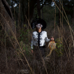 Image Detail: a photograph of a woman wearing an elaborate feathered mask wearing an black and white top and black pants holds a dead pheasant while standing in what appears to be a field overrun with brush.