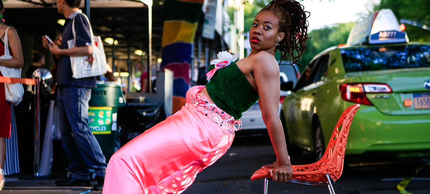 Image Detail: This is a photo of a black woman on Central Park West wearing a hot pink prom gown that has been pulled down to her waist with a piece of astro-turf wrapped around her mid section as a tube top. She is doing chair dip exercises in the street in oncoming traffic.