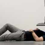 Image: Monitor installed in a gallery-like setting in an atypical spot with the screen facing the ground, only a few feet from the ground. A man lays on the floor, face up towards the screen.