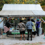 Image: a lot with a small farmstand/workspace with a garden in the background. Ten individuals are talking and interacting with each other in the phone, their action centered around the stand.