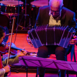 Image: Two men are seated on a stage, bathed in magenta light; one plays a violin and the other plays an accordion with a drum set in the background.