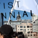 Image: A small group of artists walk together over the Brooklyn Bridge with city buildings in the background. They carry a white flag with black text that reads: "Immigrant Artist Biennial." There is a "watermark" of the Immigrant Artist Biennial logo over the image.