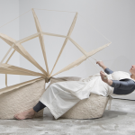 Image: In the center of the photograph is a female performer sitting on a saddle-like ceramic structure. The saddle structure is in the shape of a 2.5’ x 26’ wedge with another wedge mirroring it. We are viewing her profile. Her body position suggests movement forwards and backward while in each outstretched hands she grasps cotton twill tape which is attached to 2.5’ x 5' teardrop-shaped cloth fan blades anchored into the wedges.