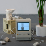 Two 1980’s era Macintosh computers are surrounded by a plaster cast polyhedron, stones, wires, cast resin Apple mice, a partial 3D print of the bust of Nefertiti, and a plastic plant.