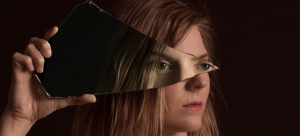 Image Detail: Close-up of a woman's face, which is turned towards the right and obscured by a shard of glass she is holding. The glass reflects a pair of eyes back, presumably the same woman's. She is dramatically lit against a black background.