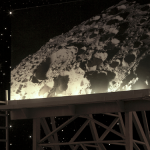 Photograph is of a large billboard, which stands against a starry night sky. On the billboard is an image of a portion of the moon, showing off one of its large craters.