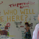 Image: A woman in a headscarf stands in the foreground with her hand on her hip, watching a young girl participating in Alexandra Chasin's "Writing On It All" project. The girl stands poised with a paintbrush, about to write a message in paint directly on a white wall, which is already covered in words and images.