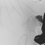 Image: Black and white photo of a girl wearing a headscarf writing on a white wall. The text begins to read: "Say What You..."