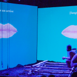 Photo of a stage with two blue screens perpendicular to each other; one says I'm an artist and has a closed mouth on it and the other says laughs also with a closed mouth on it