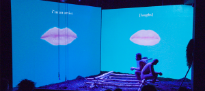Photo of a stage with two blue screens perpendicular to each other; one says I'm an artist and has a closed mouth on it and the other says laughs also with a closed mouth on it