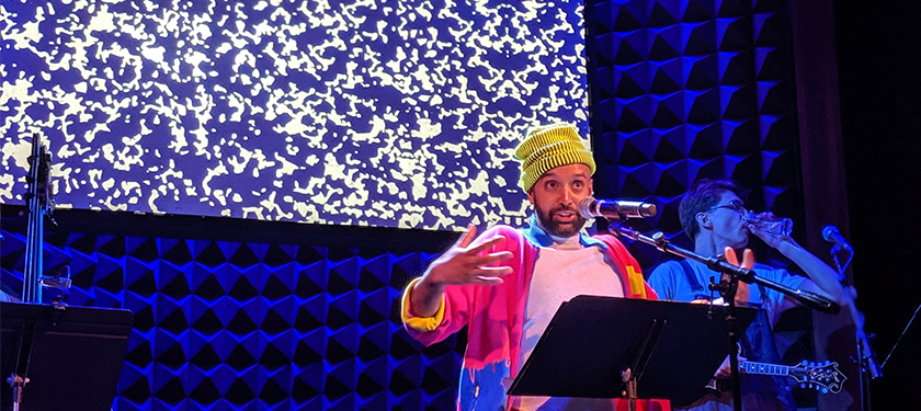 Photo of Shayok Misha Chowdhury wearing a yellow beanie and magenta jacket speaking in front of a blue textured scenery