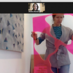 Image: Screen shot from an online studio visit being conducted by Zoom. Artist Ann Tarantino holds a bright pink piece of plastic with boldly-shaped images cut out. There is a large painting behind her on the wall. There is a small thumbnail of Heather Bhandari above.