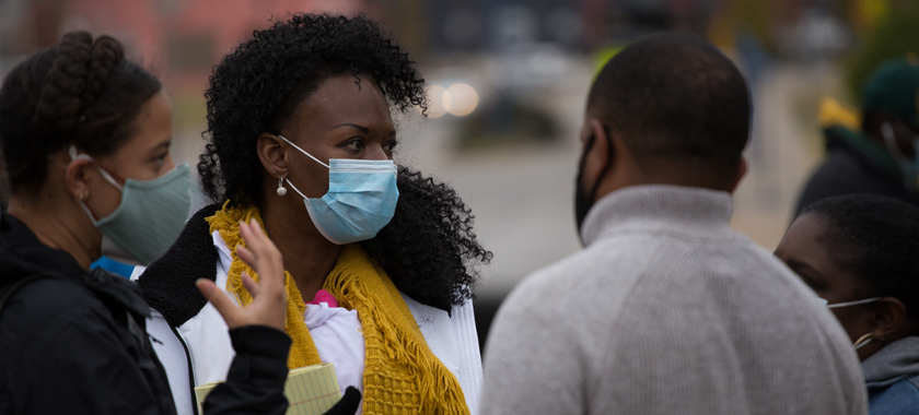 Image: A group of four people confer--all wearing masks--against a blurred background.