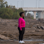 Image: A woman wearing a bright pink top, black leggings, and white sneakers stands on the bank of a river with bridges in the background.