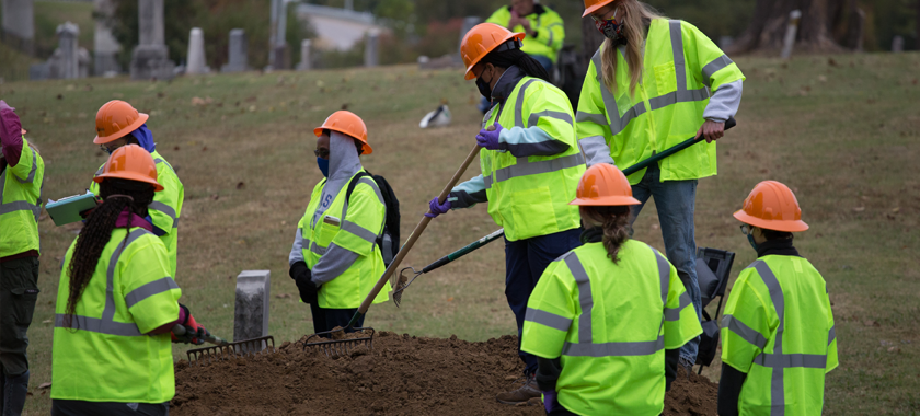 Image: A team of forensic archeologists excavate a suspected mass grave wearing bright green jackets and orange hard hats.