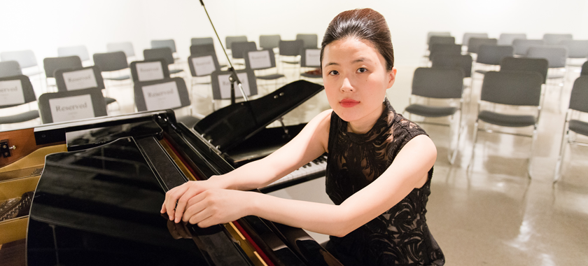 Image: Photo of Eunbi Kim at the piano in an empty performance space. Her arms are outstretched as she rests her hands on the top of the piano.