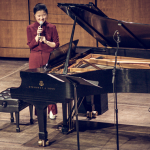 Image: Eunbi Kim stands on stage with a microphone in hand, addressing an unseen crowd at The Kennedy Center in Washington, DC.