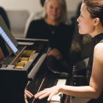 Image: Photograph of Eunbi Kim performing at Korean Cultural Center NY. She is playing two pianos at once while reading music off of an electronic device.