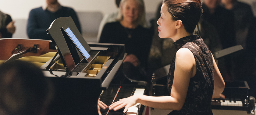 Image: Photograph of Eunbi Kim performing at Korean Cultural Center NY. She is playing two pianos at once while reading music off of an electronic device.