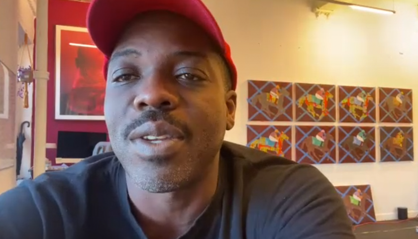 Image: a man in a black tee-shirt and red baseball cap sits in studio with artwork on the walls behind. He's pictured from the shoulders up.