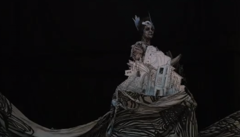 Image: Stop-motion animation still of a woman painted in black and white against a black background. She wears a cut-out of buildings on her dress, which flows into a sweeping, wave-like gesture at the bottom of the screen.