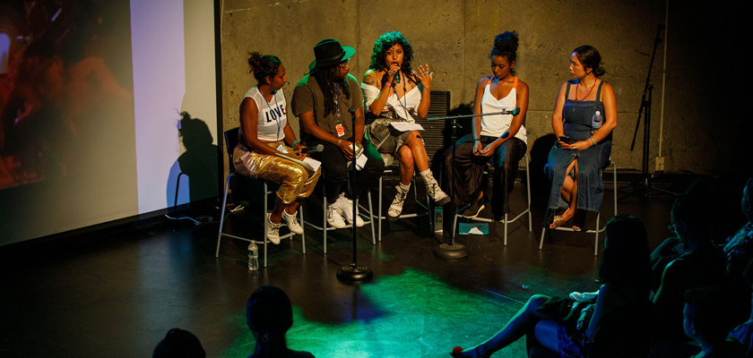 Image: Five individuals sit on raised chairs on a spotlit stage, one with a microphone in hand. A crowd sits in chairs before them.