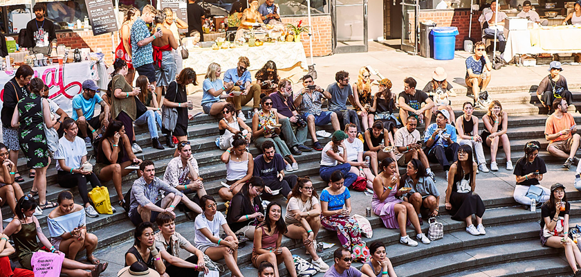 Image: A crowd gathers in the outdoor amphitheater, opposite the music stage at Happy Family Night Market 2019. Behind them are vendors seated behind tables.