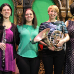 Image: Six women stand, smiling into the camera and holding woodwind and brass instruments against a colorful green backdrop.