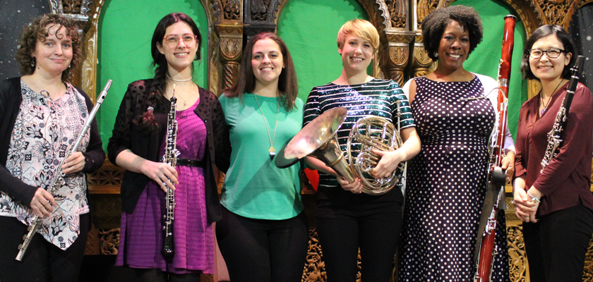 Image: Six women stand, smiling into the camera and holding woodwind and brass instruments against a colorful green backdrop.