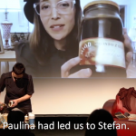 Image: A screengrab from a streaming performance-in-process. One woman is on a stage standing at a table opening a glass container. Another woman is pictured on a screen behind her, holding a glass jar filled with juice. We assume the jar they both are holding is the same.