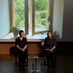 Image: Two women are seated on a stage. One is speaking, the other is silent. Behind them is a screen displaying a still photo of an open widown.