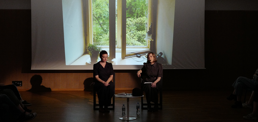 Image: Two women are seated on a stage. One is speaking, the other is silent. Behind them is a screen displaying a still photo of an open widown.