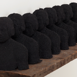 Image: Eleven busts made out of coal, lined up in a row on an aged wood-topped pedestal.