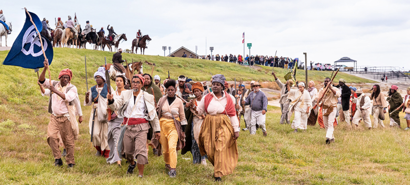 Image Detail: Dread Scott's "Slave Rebellion Reenactment," featuring contemporary people in historical garb, marching in Louisiana and armed with prop machetes, sickles, and muskets with flags flying.