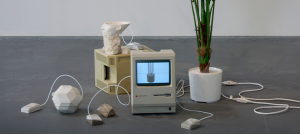 Image: An exhibition view shows a group of objects assembled on the concrete floor. Two 1980’s era Macintosh computers are surrounded by a plaster cast polyhedron, stones, wires, cast resin Apple mice, a partial 3D print of the bust of Nefertiti, and a plastic plant. The computer screens display black and white images of other plastic plants that can be purchased on the internet.